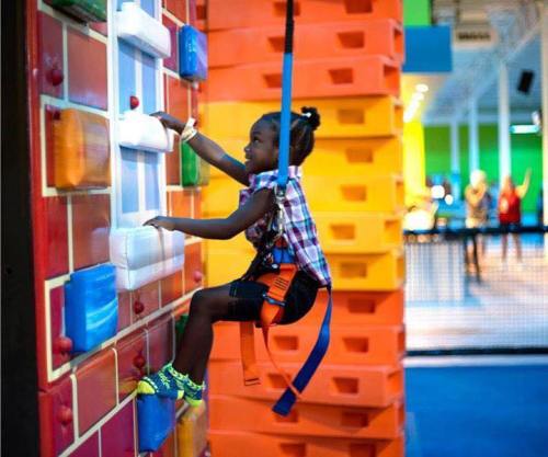 A child in a harness climbs a colorful indoor climbing wall, focused and determined.