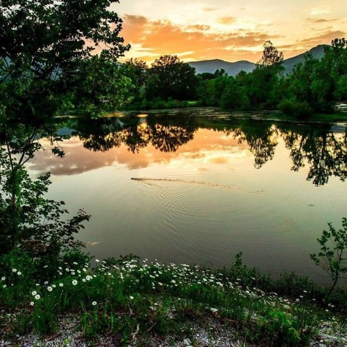 A serene sunset over a calm lake, surrounded by lush greenery and mountains, with reflections on the water's surface.