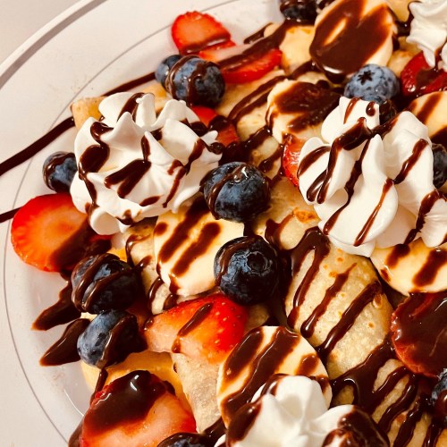 A colorful dessert plate with strawberries, blueberries, whipped cream, and chocolate drizzle on a bed of pancakes.