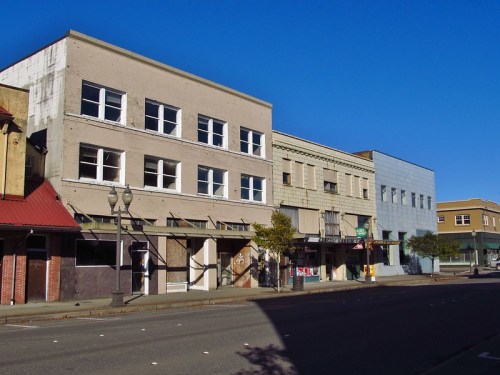 Abandoned buildings line a quiet street under a clear blue sky, showcasing faded facades and boarded windows.