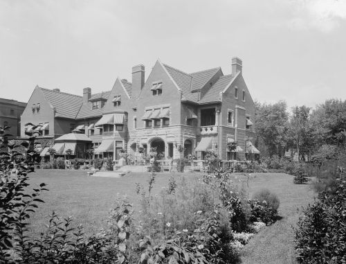 A large, historic brick mansion with a landscaped garden and patio, surrounded by trees and other buildings.