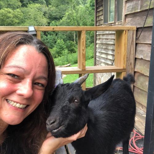 A woman smiles while taking a selfie with a black goat on a wooden porch surrounded by greenery.