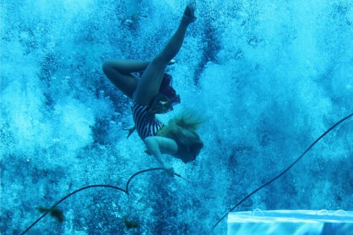 A person in a striped swimsuit is underwater, twisting and surrounded by bubbles in a blue-tinted environment.