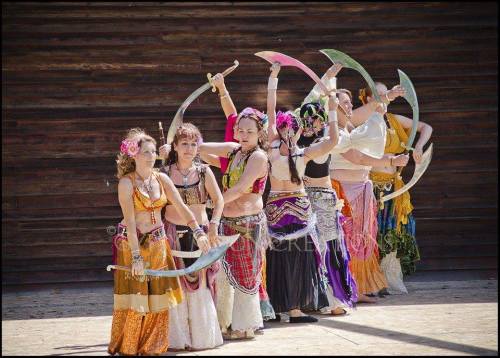 A group of dancers in colorful costumes perform with curved swords in a lively outdoor setting.