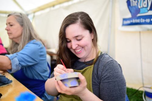 A woman smiles while painting a small bowl, with another person working in the background inside a tent.