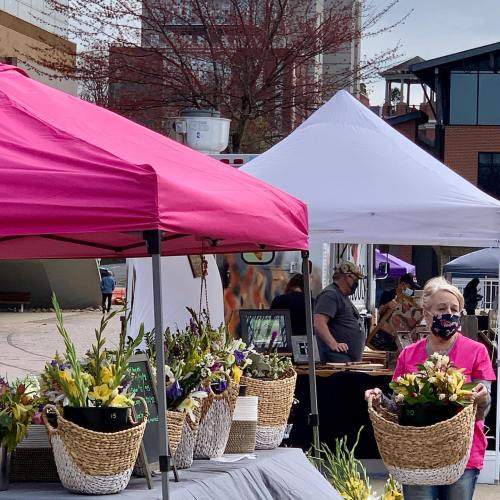 A woman in a pink shirt and mask carries a basket of flowers at an outdoor market with tents in the background.