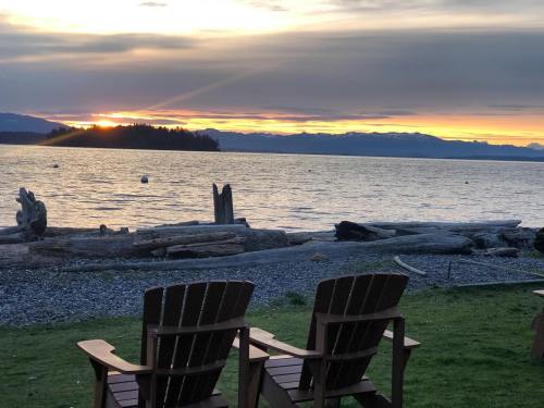 Two wooden chairs face a tranquil lake at sunset, with mountains and logs in the background.
