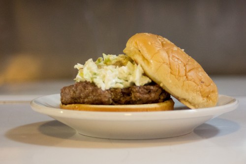 A hamburger on a plate, featuring a beef patty topped with coleslaw and served in a soft bun.
