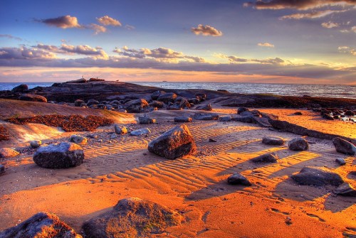 A rocky beach at sunset, with golden sand patterns and clouds reflecting warm colors in the sky.