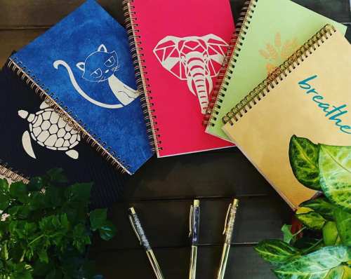 A collection of colorful spiral notebooks with animal designs, surrounded by green plants and pens on a wooden surface.