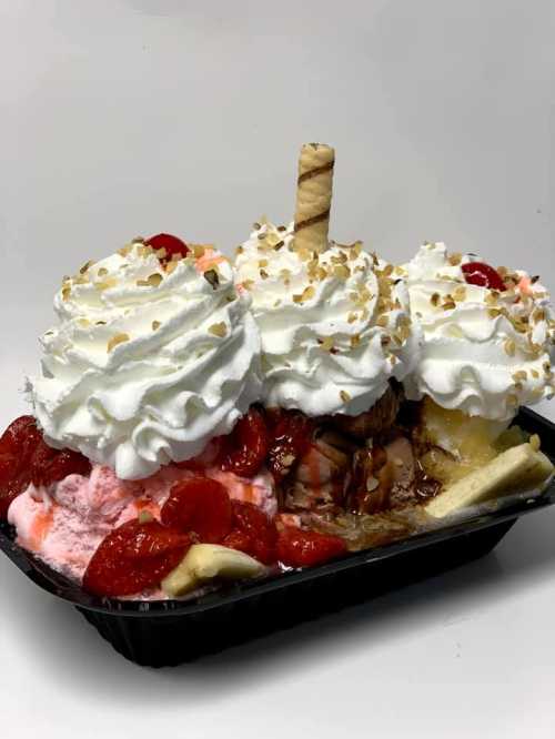 A colorful ice cream sundae with whipped cream, strawberries, and a wafer stick, served in a black container.