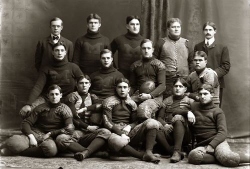 A vintage black-and-white photo of a football team, featuring 12 players and 3 coaches in formal attire.