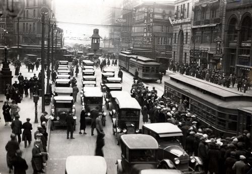 A busy city street in the early 20th century, filled with cars, streetcars, and crowds of people.