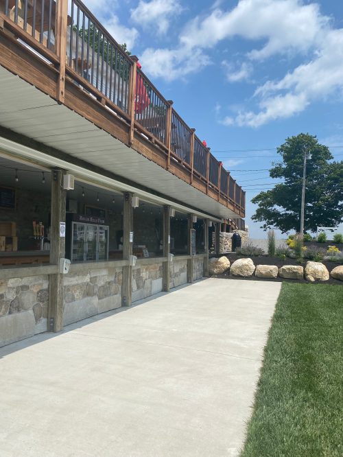 A wooden deck overlooks a stone patio and grassy area, with a bar and trees in the background under a blue sky.