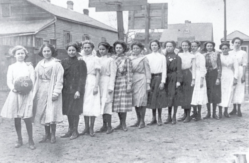 A historic black-and-white photo of a line of young women in early 20th-century clothing, standing outdoors.