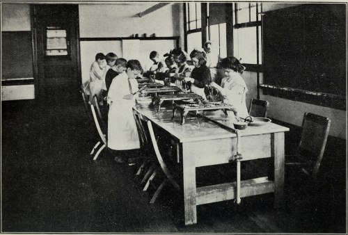 Students in a classroom, focused on a long table with various tools and materials, engaged in hands-on activities.