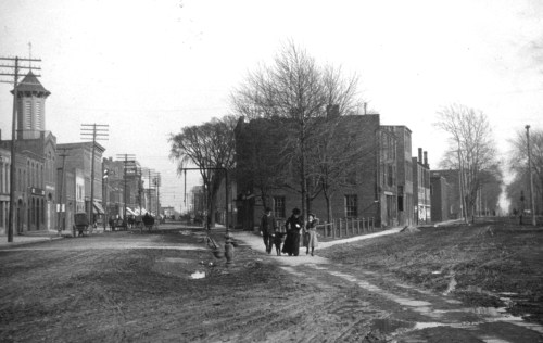 Black and white photo of a dirt street with buildings, trees, and people in early 20th-century urban setting.