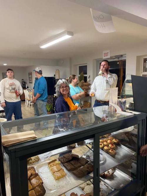 A group of people socializing in a bakery, with a display case of baked goods in the foreground.