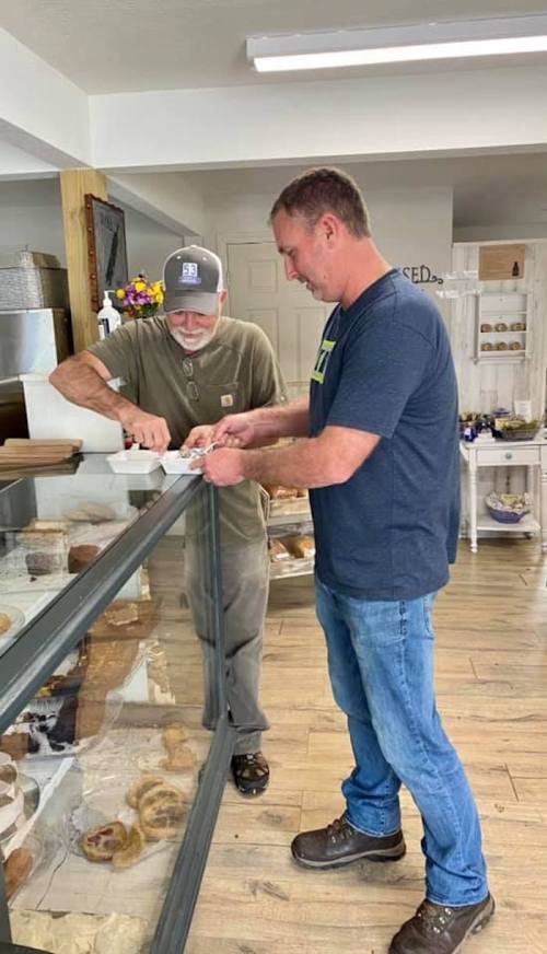 Two men in a bakery, one serving pastries from a display case while the other looks on, engaged in conversation.