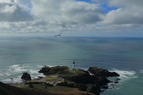 A scenic view of a lighthouse on a rocky coast, with a plane flying above and a calm ocean under a cloudy sky.