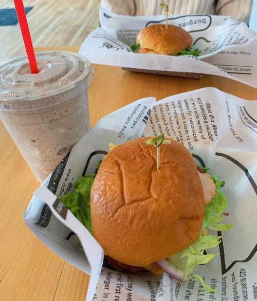 A close-up of a sandwich with lettuce on a paper wrapper, alongside a milkshake in a cup with a red straw.