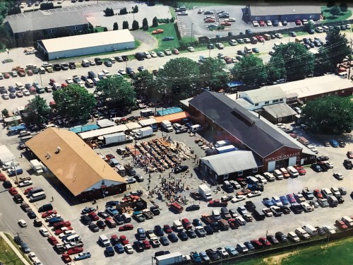 Aerial view of a busy outdoor market with many cars parked and people gathered near a large building.