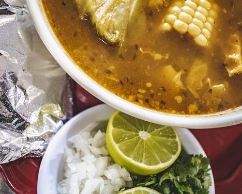 A bowl of soup with corn and chicken, accompanied by lime, chopped onions, and cilantro on the side.