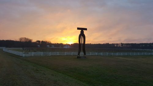 A large sculpture stands in a field at sunset, with a white fence and trees in the background.