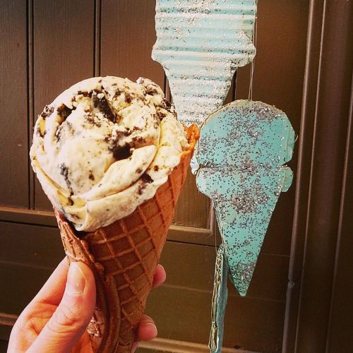A hand holding a cone of cookies and cream ice cream next to a decorative ice cream sign.