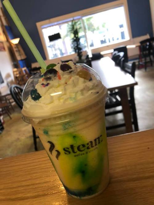A colorful layered drink with whipped cream and toppings, served in a clear cup with a green straw at a café.