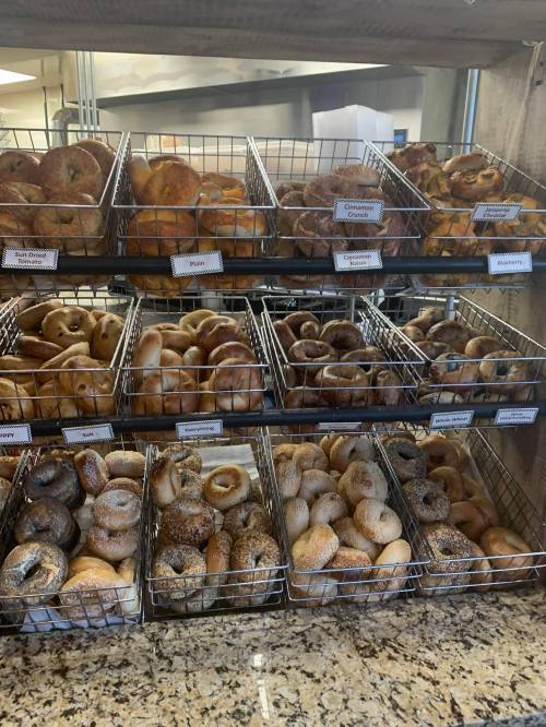 A display of various bagels in wire baskets, labeled with different flavors, on a countertop.