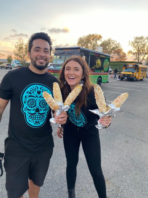 A man and woman hold corn on the cob, smiling, wearing matching black shirts with a blue skull design. Food trucks in background.
