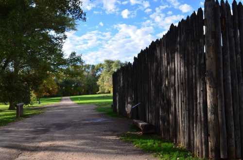 A dirt path leads through a grassy area, bordered by a tall wooden fence under a blue sky with scattered clouds.