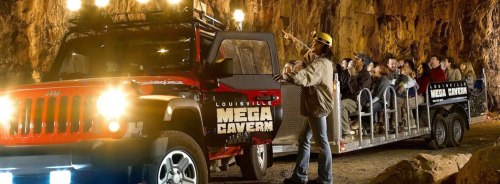 A tour guide leads a group on a tram inside a cavern, with a red vehicle parked nearby, illuminated by overhead lights.