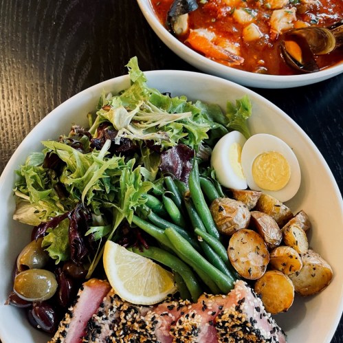 A bowl of salad with greens, green beans, potatoes, sesame-crusted fish, and a lemon wedge, alongside a bowl of seafood stew.