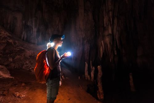 A person with a backpack holds a flashlight, exploring a dark cave with stalactites in the background.