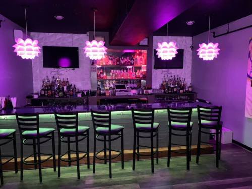 A modern bar with purple lighting, high stools, and a well-stocked shelf of liquor in the background.