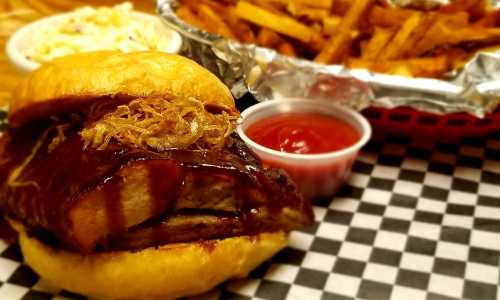 A close-up of a BBQ sandwich with pulled meat, topped with onions, served with fries and dipping sauce on a checkered table.