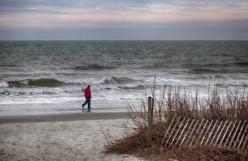 A person in a red jacket walks along a sandy beach with waves and a cloudy sky in the background.