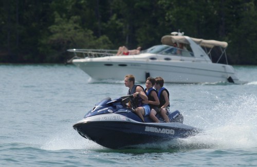 Three people ride a jet ski on a lake, with a boat in the background and trees lining the shore.
