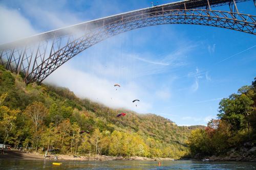 Paragliders soar beneath a large bridge over a river, surrounded by colorful autumn trees and a clear blue sky.