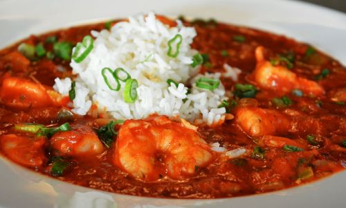 A bowl of shrimp in a spicy red sauce, topped with green onions and served with a mound of white rice.