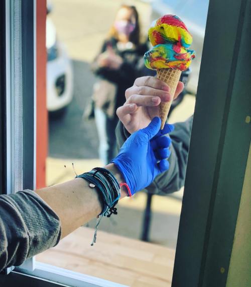 A hand in a blue glove hands a colorful ice cream cone to another hand through a window.