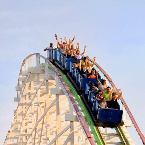 A group of excited riders on a roller coaster, hands raised, descending a steep track against a clear sky.