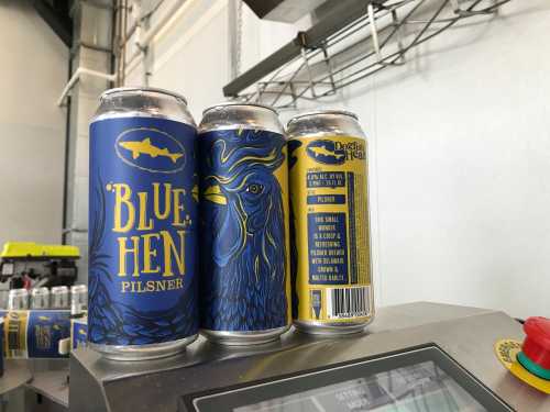 Three cans of "Blue Hen Pilsner" featuring a blue and yellow design with a rooster illustration.