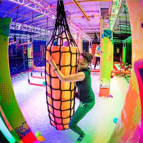 A person climbs a colorful, netted obstacle in a vibrant indoor adventure park.