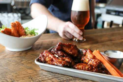 A platter of glazed chicken wings with carrots and dipping sauce, alongside a bowl of salad and a beer in the background.