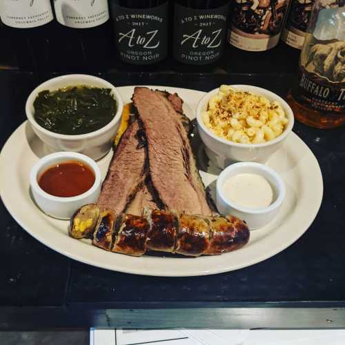 A plate featuring brisket, sausage, macaroni and cheese, collard greens, and dipping sauces.