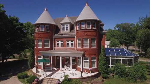 A large, red brick house with turreted towers, a wraparound porch, and a glass sunroom surrounded by greenery.