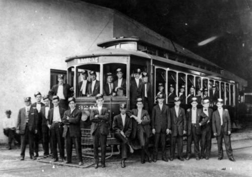 Historic black-and-white photo of a streetcar with a large group of men, likely workers, posing in front of it.